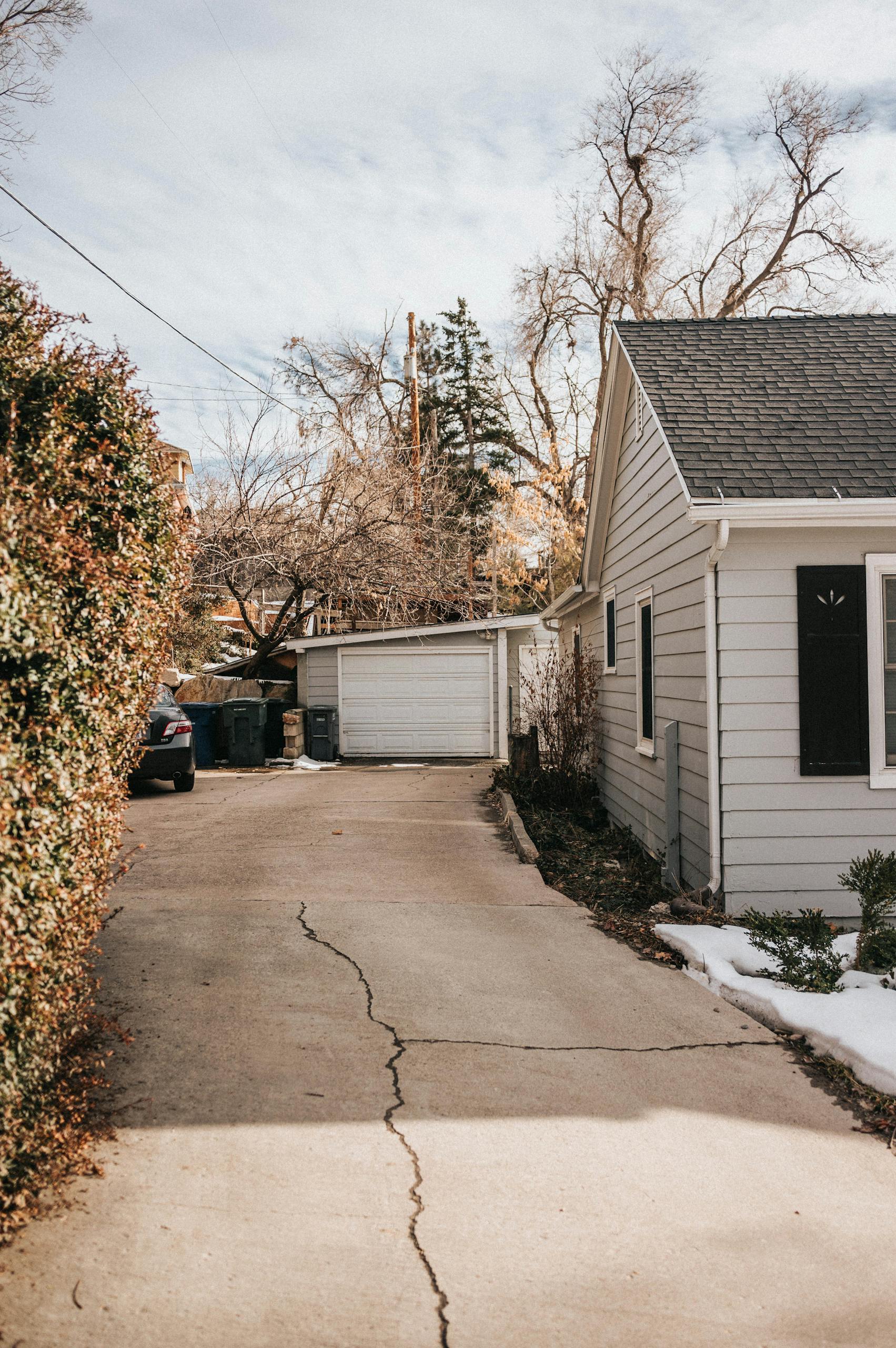A peaceful suburban driveway with cracked pavement leading to a house surrounded by bare trees.